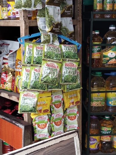 A cluttered display features an assortment of food items. Packages of "Wonda Thyme Leaves" and "Marico Dry Rosemary Leaves" hang from a blue hanger. Below these are packets of "Gino Peppered Chicken" and "Addme" products. To the left, there are bags of rice, "Big Bull" snacks, and "G&SEL Basmati." On the right, a shelf holds various brands of cooking oil, including "Power Oil," "Morga," "Golden Penny Pure Soya Oil," and "Namador."