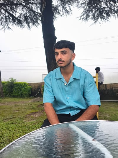 A young man with dark, short hair and a mustache sits on a golden chair, facing slightly away from the camera. He is wearing a turquoise and white striped short-sleeved collared shirt and black pants, with a silver chain around his neck. Behind him, a second man in a white shirt stands near a low stone wall. The scene appears to be an outdoor seating area, possibly a cafe or restaurant, with a glass-topped table in the foreground. Lush green grass and trees, including a large tree with intricate branches, surround the area. The background is dominated by a pale, overcast sky and power lines. The location is identified as Khaniara, India.