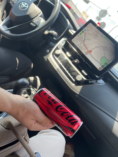 The image shows a person holding a can of Coca-Cola Zero Sugar inside a car. The car's interior is visible, including the steering wheel with the Toyota logo, the dashboard with a navigation screen displaying a map, and the center console with a gear shift. The navigation screen shows a route with traffic information. A person's hand is holding the red and black can of Coca-Cola Zero Sugar. In the background, a person's legs are visible, wearing light-colored pants. The overall scene suggests the person is either driving or a passenger in the car. The lighting indicates it is daytime, with sunlight casting shadows inside the car. The scene is set within a car, with the dashboard and steering wheel prominently featured. The text "Coca-Cola", "ZERO SUGAR", and "NO CALORIES" are visible on the can. The location is identified as Katowice, Poland.