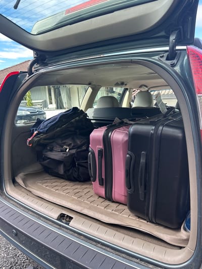 The image shows the open trunk of a dark green car, packed with luggage. Two suitcases, one black and one pink, are stacked vertically. The black suitcase is on top and has a black handle. The pink suitcase is below it and also has a black handle. There is a black backpack and a pile of dark clothing placed in front of the suitcases. The car interior appears to be a light gray fabric. The trunk floor is a textured beige plastic. The scene is viewed from above, looking into the trunk. Outside the trunk, there is a hint of a building with a gray roof and white siding. The sky is visible in the background, showing blue with some white clouds. The lighting suggests it is daytime. No people are visible. The location context is Kongsberg, Norway. No visible text is apparent in the image.