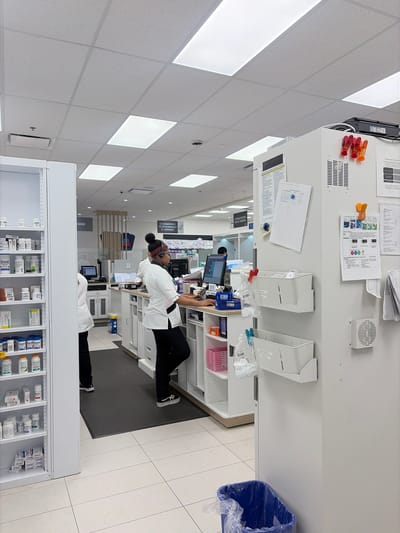 This is a medium-shot, eye-level, indoor photograph of the interior of a pharmacy. The scene depicts a checkout counter area with two pharmacy technicians working. A pharmacy technician, wearing a white uniform and headset, stands behind the counter, facing right, and appears to be operating a computer. Another pharmacy technician, also in a white uniform, stands to the left, partially obscured by a shelving unit, and is facing the counter.

The pharmacy is well-lit with fluorescent ceiling lights and has a clean, modern aesthetic. White shelving units line the walls, filled with various pharmaceutical products. A blue trash can is visible in the lower right corner of the image.

The location is identified as a pharmacy in Longueuil, Canada. This is evident from the signage within the pharmacy, which includes French text, a common feature in Quebec. For example, the sign above the checkout counter reads "CAISSE", which is French for "cashier" or "checkout". Another sign visible says "COMMANDES TÉLÉPHONIQUES ET INTERNET", meaning "phone and internet orders". The overall environment suggests a commercial setting within a retail establishment.