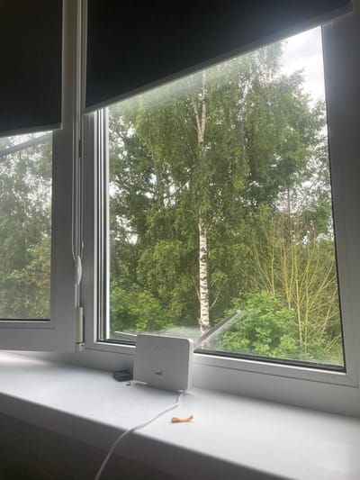 The image displays a view from a window of a residential building, likely in Коробицыно, Russia, overlooking a green, wooded area. The window features white frames and roller blinds, one of which is partially drawn. On the white windowsill, a white wireless router with four indicator lights and "4G+5G" branding is visible. A black rectangular object, possibly a power adapter, and a small piece of orange plastic are also present on the windowsill, along with a white power cord connected to the router. The outdoor scene beyond the window is dominated by lush green foliage, including the distinct white bark of a birch tree and other deciduous trees and shrubs. The sky appears overcast.