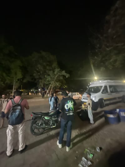 A nighttime scene captures a group of people near a white van, parked on a paved surface with some vegetation and trees in the background. In the foreground, a person wearing a pink shirt and a backpack faces away from the camera, walking towards the right. Next to them is a dark-colored motorcycle. To the right of the motorcycle, another person in a dark t-shirt and blue jeans also faces away from the camera. A white van is parked to the right, with a person in a light-colored outfit standing near it, appearing to be loading or unloading luggage. Two large blue bins are situated behind the van. The ground shows scattered debris, including what appears to be plastic bottles and wrappers. The overall lighting suggests artificial illumination from streetlights or vehicle headlights. The scene appears to be an outdoor area, possibly a parking lot or a roadside in Kajheri, India.