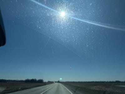 The image is a view from inside a vehicle looking out through the windshield. The windshield is dirty with visible water spots and streaks. A crack is present in the windshield, curving across the middle of the frame. The sun is bright and high in the sky, causing glare. Outside the vehicle, a straight paved road stretches into the distance, flanked by flat fields and sparse trees on either side. There is no indication that the location is in the city of Обухово, Russia. A small insect is stuck to the windshield, near the center of the frame. No people are visible.