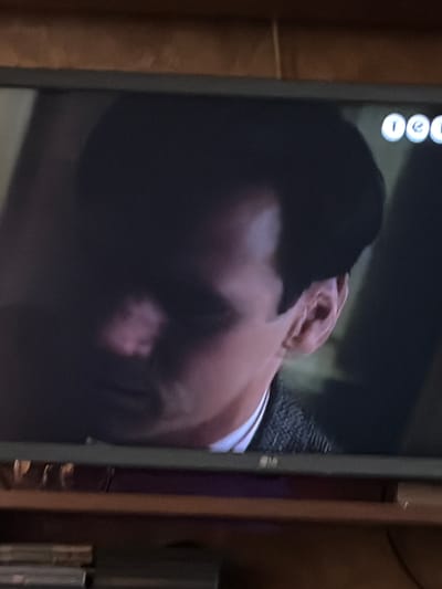 The image shows a close-up of a television screen displaying a man's face. The man has dark hair and is looking off to the side. His face is partially obscured by shadow, but his ear and the side of his head are visible. He is wearing a dark jacket over a white shirt. The television screen is mounted on a shelf, and the top of the shelf and a portion of the wall are visible in the background. There are some circular icons in the upper right corner of the television screen. The image is grainy and out of focus, suggesting it was taken with a camera or phone from a distance. There is no identifiable information in the image that would suggest a location in Osolynka, Ukraine.