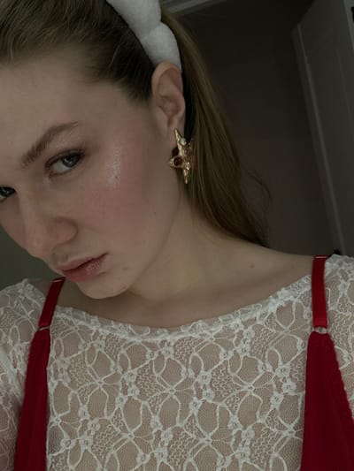 A woman's face, angled slightly, features a gold, multi-colored gemstone earring shaped like a star. A white headband sits atop her blonde hair. Her skin is fair, with a hint of pink in her cheeks. She wears a white lace garment with red straps. The background is muted and dark.