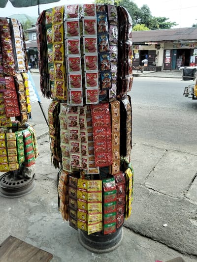 Two cylindrical displays showcase various products, mainly food seasoning packets. The larger display features packets with labels such as "K", "ARSOR", and "Wanda", arranged in rows around the cylinder's perimeter. The smaller display exhibits similar packets, including "BENNY", arranged similarly. These displays stand on circular metal bases, positioned on a concrete surface near a street. The background reveals a street scene with buildings and some people.