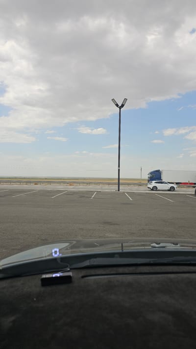 The image captures a view from inside a vehicle, looking out at a parking lot and a distant landscape under a cloudy sky. A dual-headed street lamp stands prominently in the middle ground. In the background, a blue truck and a white SUV are visible on a road bordering a vast, flat, arid area. The parking lot is paved and marked with white lines. The interior of the vehicle shows a portion of the dashboard with a head-up display unit projecting a digital readout onto the windshield. The location is identified as Raz'ezd Kaynar, Kazakhstan.