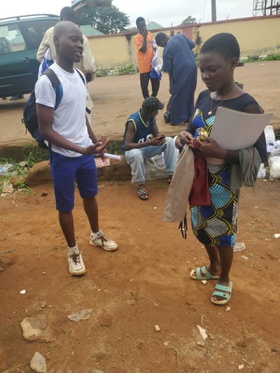 In the city of Bahit, Nigeria, two young people, a male and a female, stand on a dirt road. The male, facing left and smiling, wears a white t-shirt, blue shorts, and white sneakers. He has a backpack slung over his left shoulder and is holding a small piece of paper. The female, facing forward and looking down, wears a black top with a colorful, patterned skirt. She holds a clipboard and a bundle of cloth items. In the background, other individuals are visible, some near a green vehicle and a yellow wall. The environment appears to be outdoors, with scattered debris on the ground.