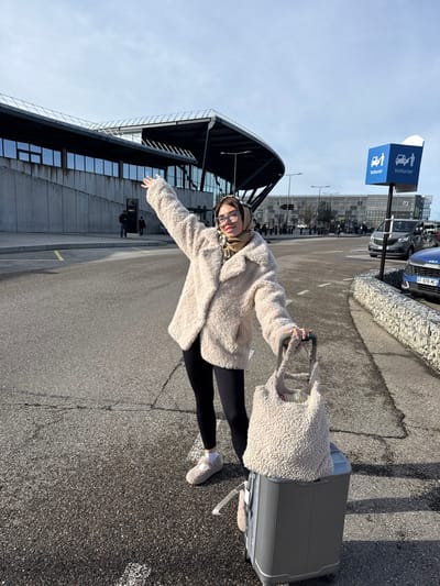 A female individual stands on asphalt pavement at Lyon-Saint Exupéry Airport in Colombier-Saugnieu, France. She is wearing glasses, a light brown patterned headscarf, a cream-colored fuzzy fleece jacket, black ribbed leggings, and light-colored fuzzy slippers with white socks. She smiles and looks directly at the camera, her right arm raised and extended outwards, while her left hand grips the handle of a grey hard-shell rolling suitcase. A cream-colored fuzzy tote bag, matching her jacket, rests on top of the suitcase. In the background to the left, a modern airport terminal building features a dark, curved roof, large horizontal glass windows, and a concrete base. To the right, a blue sign displays two white pictograms and the text "Voiturier" (valet), with a "P3" sign visible behind it. Multiple vehicles, including a dark grey van and a partially visible blue car (license plate GT 978 MC), are parked or moving further down a road. The scene is under bright daylight with a partly cloudy sky.
