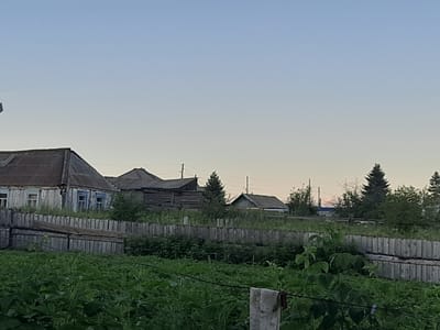 The image depicts a rural scene in the city of Kamarchaga, Russia. A weathered, wooden house with blue-trimmed windows and a corrugated metal roof is visible on the left side of the frame. A wooden fence runs horizontally across the midground, separating a cultivated area from the foreground. Beyond the fence, several other wooden structures and trees are scattered throughout the landscape under a pale sky. The foreground is dominated by lush green vegetation, with some plants taller than others. A clothesline with a piece of fabric hanging from it is also present in the foreground. The overall impression is one of a quiet, somewhat dilapidated, rural settlement.