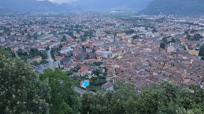 This is an aerial, high-angle, wide shot of the town of Riva del Garda, Italy, taken at dusk. The foreground is dominated by lush green trees, with branches and leaves obscuring the lower portion of the image. Beyond the foliage, the dense urban landscape of Riva del Garda unfolds. Buildings with terracotta tile roofs and pastel-colored facades are clustered together, forming a characteristic Mediterranean townscape. Streets wind through the town, with faint traces of car headlights visible. In the mid-ground, a bright blue swimming pool stands out amidst a green, manicured area, possibly a hotel or residential complex. The background shows the distant, hazy outlines of mountains shrouded in the twilight. The overall impression is of a peaceful, densely populated town nestled between verdant hills and dramatic mountain scenery.