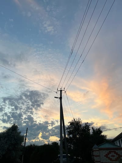 The image depicts a rural scene in Kotovo, Russia, at sunset. The sky is filled with a mixture of blue, gray, and pink clouds, indicating the transition from day to night. Several power lines stretch diagonally across the frame, supported by a wooden utility pole in the foreground. Silhouetted against the colorful sky are dark green trees and the roofline of a building with a brick wall. The overall atmosphere is serene, with the natural beauty of the sunset contrasting with the utilitarian presence of the power lines and man-made structures.
