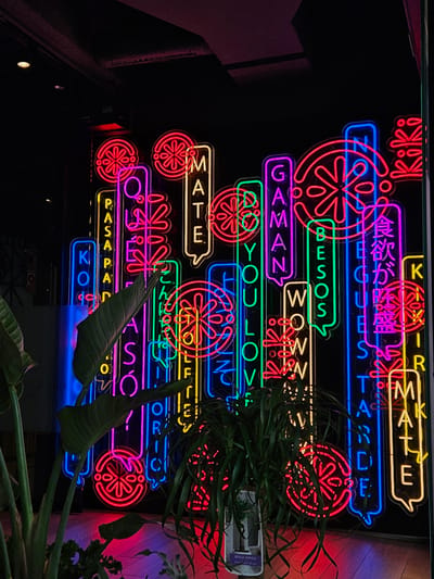 This image features a vibrant and eclectic display of multicolored neon signs illuminating a dark indoor setting. Various neon tubes in red, yellow, green, blue, purple, and pink create a dynamic visual tapestry of text and decorative red circular patterns resembling stylized fruit slices or abstract flowers. The signs are arranged predominantly horizontally, often framed within speech bubble-like outlines.
Visible text on the neon signs includes a mix of languages. English phrases such as "Kiki," "KIKIRMATE," "SMATE," "WOWWW," "GAMAN," "DO YOU LOVF," and "MATE" are prominently displayed. A Spanish word, "BESOS" (kisses), is rendered in green neon. Japanese characters for "食欲が黒" (appetite is black) appear in pink. Other text includes "EGUES TANDE," "PASAPAN," and "KO."
Several green plants with broad leaves and narrower foliage are interspersed throughout the scene, partially obscuring some of the neon elements. A white label on a container behind one plant clearly reads "BEAUCARNEA," identifying it as a type of ponytail palm. The dark background emphasizes the glowing lights, suggesting a nighttime ambiance or a dimly lit interior. The image, taken at an upward angle, conveys the energetic and modern atmosphere of what appears to be a trendy bar, restaurant, or lounge in Santa Cruz De Tenerife, Spain.