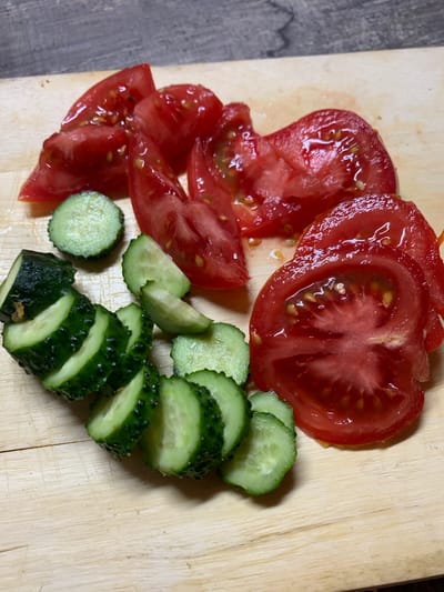 This overhead close-up shot captures a vibrant arrangement of freshly sliced red tomatoes and green cucumbers, presented on a light wooden surface. The tomatoes, some in full round slices and others in wedges, appear juicy with visible seeds and liquid glistening on their bright red flesh. Adjacent to them, a pile of neatly sliced cucumbers exhibits their dark green, textured skin and pale green interior, looking crisp and fresh. The wooden surface, possibly a cutting board, shows some minor imperfections and a light grain. A darker, textured surface, likely a countertop or table, is partially visible in the left background. The image depicts the preparation of fresh, healthy vegetables, ready to be consumed or incorporated into a dish. No people or text are visible in the frame. The scene is set in Kasimovo-1, Russia.