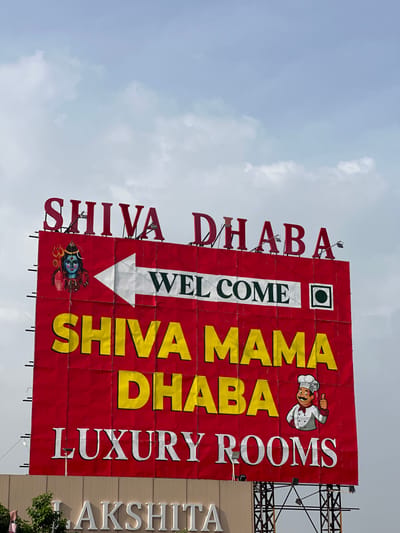A billboard for "SHIVA MAMA DHABA LUXURY ROOMS" is pictured against a cloudy sky in Dhani Gaiskan, India. The red billboard features large, red lettering at the top spelling "SHIVA DHABA". Below this, a white arrow points to the right, superimposed over the words "WELCOME" in green text. To the left of the arrow is an image of Lord Shiva. The lower half of the billboard displays "SHIVA MAMA DHABA" in large yellow letters. To the right of this, a cartoon chef character gives a thumbs-up. The words "LUXURY ROOMS" are written in white at the bottom of the red portion. Below the red billboard, a beige structure with white lettering spelling "AKSHITA" is partially visible. The scene is set outdoors, with trees and a metal frame supporting the billboard visible at the bottom.