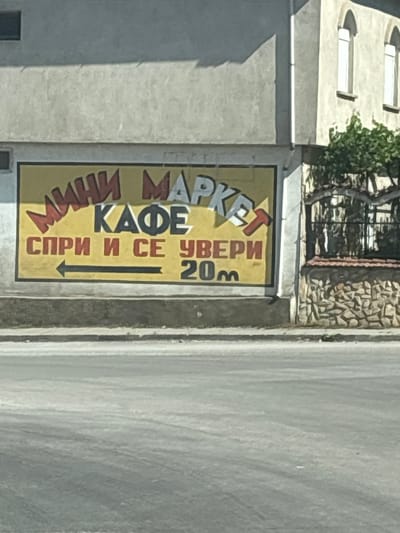 The image shows a sign on the exterior of a building, likely in Topolovgrad, Bulgaria, advertising a "MINI MARKET KAFE". The sign is yellow with red and white lettering. The top line reads "МИНИ МАРКЕТ" (MINI MARKET) in red and white. The second line says "КАФЕ" (CAFE) in black. The third line reads "СПРИ И СЕ УВЕРИ" (STOP AND SEE FOR YOURSELF) in red, with a black arrow pointing left. Below this, "20m" is written in black. The building appears to be made of concrete. To the right of the sign, there is a stone wall topped with a wrought-iron fence and some greenery. The scene is viewed from across a paved road, suggesting it is a roadside establishment.