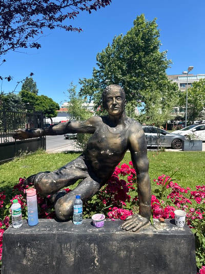 This is a medium-shot, eye-level outdoor photograph taken on a bright, sunny day in Sancaktepe, Turkey. The primary subject is a bronze statue of a man in a dynamic, athletic pose, possibly mid-stride or about to leap. He is leaning forward with his left arm extended forward and slightly upward, while his right arm is bent at the elbow and resting on the dark, rectangular base of the statue. The statue is weathered and shows patches of lighter color on its dark bronze surface.

The statue is situated in front of a vibrant bed of pink roses, with green grass and other foliage surrounding it. Behind the statue, a black metal fence is visible, and beyond that, a street with parked cars, including a light-colored sedan and a white hatchback. In the background, there are trees and a multi-story building with windows. The sky is a clear, bright blue.

On the dark base of the statue, to the left of the statue's legs, are four objects: a small bottle of water with a green label, a tall, pink and blue water bottle, another bottle of water with a blue label, and a small purple container. To the right of the statue's right hand, there is a white paper cup with writing and a small dark object, possibly a seed or a pebble. The overall scene suggests an urban park or public space.