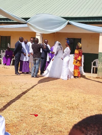 The scene depicts an outdoor gathering, likely a wedding ceremony, in Angwan Sarki, Nigeria, in front of a single-story building. The building features a light beige facade, two dark rectangular doors on the left, and a dark green corrugated metal roof. A light blue fabric canopy is draped overhead, suspended from the building. The ground is dry, light brown earth with sparse patches of dried grass, and a prominent diagonal shadow extends across it. A small red object lies on the ground in the foreground.
Ten individuals are present. In the center-right, a bride wears a white wedding gown and veil, standing next to a man in a dark suit who has his left arm extended towards her. To their right, a woman in a white long-sleeved lace dress with a dark blue headwrap stands beside a woman in a vibrant red and gold patterned traditional outfit with a yellow headwrap.
To the center-left, a man in a dark patterned short-sleeved shirt and blue jeans holds a device, possibly a camera or phone, with both hands raised, appearing to photograph or video the event. Behind him are three men: one in a dark suit, one in a light blue robe, and one in a white shirt with sunglasses. On the far left, two individuals in flowing purple robes are seated on white plastic chairs. Another empty white plastic chair is visible on the far right.