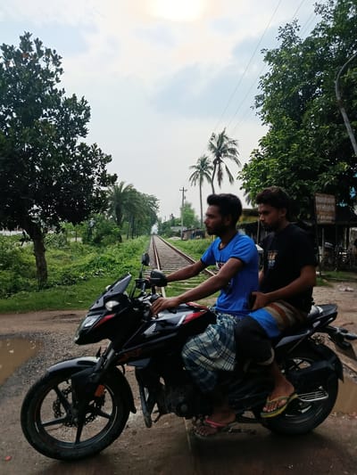 Two young men are riding on a black motorcycle on a dirt road beside a railway track in Benapole, Bangladesh. The man in the front is driving and wearing a blue t-shirt and a checkered lungi. The man in the back is wearing a black t-shirt and striped shorts. The motorcycle is positioned on the left side of the image, facing forward. The railway track runs diagonally from the middle to the right side of the image. Lush green vegetation lines both sides of the railway track, with trees and palm trees visible. The sky is overcast with some patches of sunlight. There are puddles of water on the dirt road in the foreground. A small building with a sign in Bengali is visible on the right side of the image, near the railway track. The overall scene depicts a rural or semi-urban setting in Bangladesh.