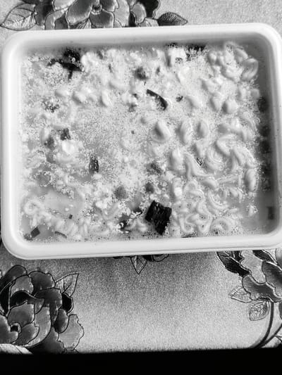 A top-down, black and white, medium shot shows a white rectangular tray filled with prepared instant noodles in broth. The noodles are coiled and submerged in a foamy, light-colored liquid with visible flecks of dark ingredients. The tray is positioned on a textured surface with a floral pattern. The background is dark, suggesting it is a tabletop or counter. There are no people or animals visible in the image. The location in the city of Dubovoye, Russia, is not discernible from the image itself.