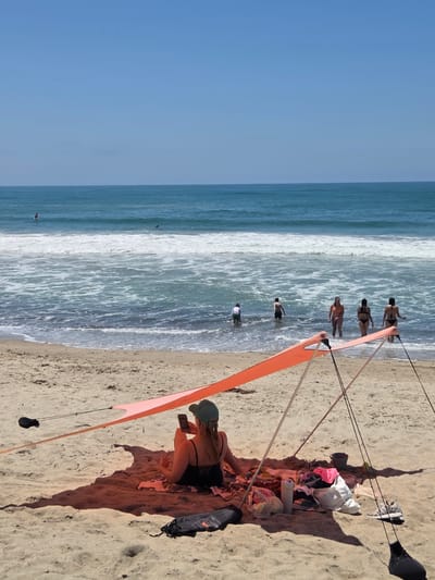 The image depicts a sunny day at a beach, likely in San Clemente, California, with the Pacific Ocean in the background. In the foreground, a person is sitting on a towel under a small orange beach tent, looking at their phone. Scattered around them are beach accessories including a water bottle, a bag of items, and a pair of sneakers. Further out in the ocean, several people are visible wading in the water and some are in the process of surfing. The sand is light brown, and the water is a clear blue with white waves. The sky is a bright, cloudless blue.