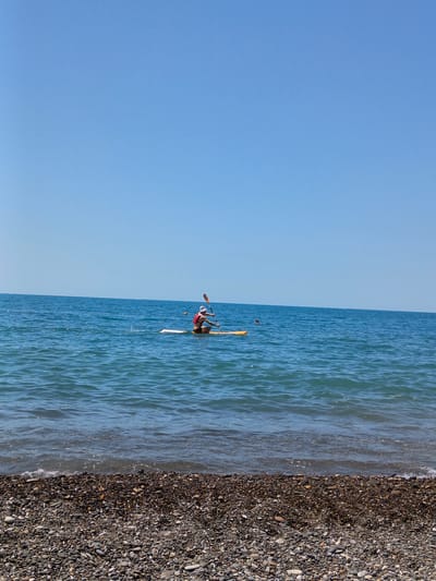 In Krinitsa, Russia, a person is paddleboarding in the clear blue sea. The individual is seated on the paddleboard, wearing a red life vest and a white hat, and is holding a paddle, actively paddling. The paddleboard is yellow and white. There are small waves in the water. In the background, the sky is clear and blue, meeting the horizon of the sea. The foreground of the image shows a pebbled beach.