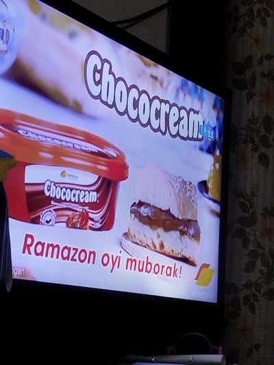 This image captures an angled, close-up view of a television screen displaying an advertisement, located within an indoor setting in the city of Chumbagish, Uzbekistan. The advertisement features a chocolate spread product named "Chococream," shown as a red rectangular tub and as a spread filling a horizontally sliced piece of light-colored bread or cake. The word "Chococream" is prominently displayed in large white letters with a brown outline across the upper portion of the screen. Below the product display, the Uzbek phrase "Ramazon oyi muborak!" (Happy Ramadan month!) is visible in red text, alongside a "N'MEDOV" brand logo in the lower right corner. A digital time indicator showing "18:22" is present in the upper right quadrant of the screen.
To the right of the television, a wall is visible, covered in patterned wallpaper featuring light brown floral or leafy designs on a lighter background. A black electrical cable is discernible above the television. The bottom left corner of the frame is partially obscured by a blurry object, possibly a person's arm or another item, exhibiting yellow and blue colors. Below the television, indistinct objects, including what appears to be a pair of pink-framed glasses and a white rolled item, rest on a surface. No individuals are clearly identifiable, and no explicit actions or interactions beyond the static display of the advertisement are depicted.