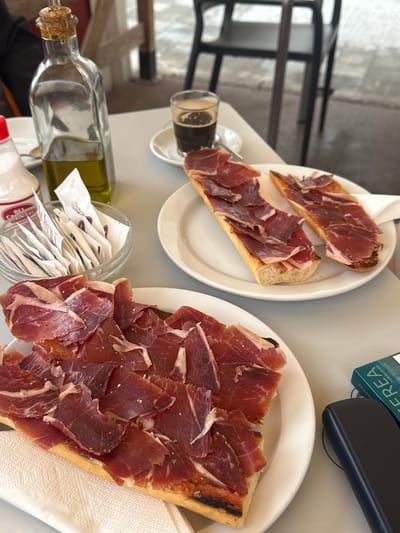 The image depicts a table setting in a restaurant, likely outdoors, in Llucmajor, Spain. Two white plates display "pan con jamón", bread topped with Spanish cured ham. A small glass of dark liquid, potentially coffee or beer, sits near one plate. An open glass container holds many small, individually wrapped items. An olive oil bottle and a bottle of mayonnaise are also on the table. A pack of cigarettes and a black phone are also sitting on the table.