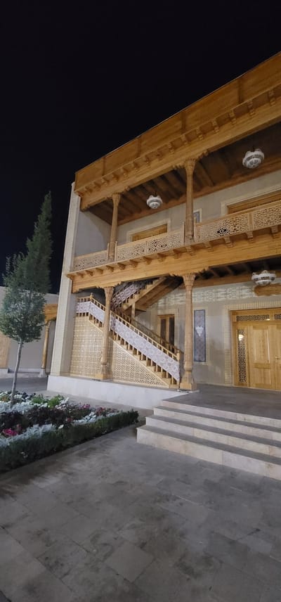The image depicts a traditional wooden building at night in Satkak, Uzbekistan. The building features intricately carved wooden balconies, columns, and decorative railings. A prominent wooden staircase with carved balustrades ascends to the upper levels. The walls of the building are made of light-colored brick, with decorative patterns. In front of the building, there is a paved courtyard with a flowerbed containing various colorful flowers and foliage. A tall, slender tree stands to the left of the building, and a set of stone steps leads into the courtyard from the right. The scene is illuminated by overhead lighting, casting soft light on the architecture and surroundings. The sky above is dark and clear.