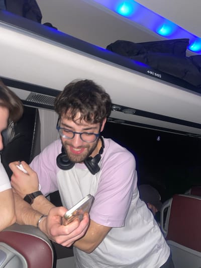 A young man with brown hair and glasses is on a bus or airplane. He is wearing a light pink shirt and has headphones around his neck. He is looking at his phone and smiling. Another person is visible in the bottom of the image. Overhead compartments and overhead lights are visible.