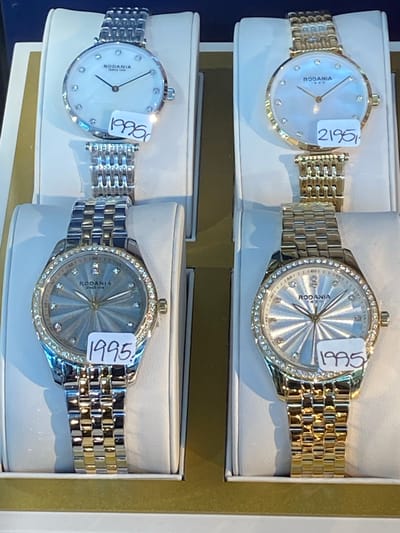 The image shows a display of four watches in a retail setting, possibly a jewelry store. The watches are arranged in pairs, with two watches on the top row and two on the bottom row. All watches appear to be of a similar style, with round faces and metal bands. The top row features two watches with silver-toned bands and white faces, one with a price tag of "1995" and the other with a price tag of "2195". The bottom row displays two watches, both with diamond-encrusted bezels. The watch on the left has a silver and gold-toned band and a blue-grey face with a price tag of "1995". The watch on the right has a gold-toned band and a white face, with a price tag of "1995". The watches are presented on white display stands within a box-like structure. The overall setting suggests a commercial environment. The location of Herning, Denmark is not discernible from the image.