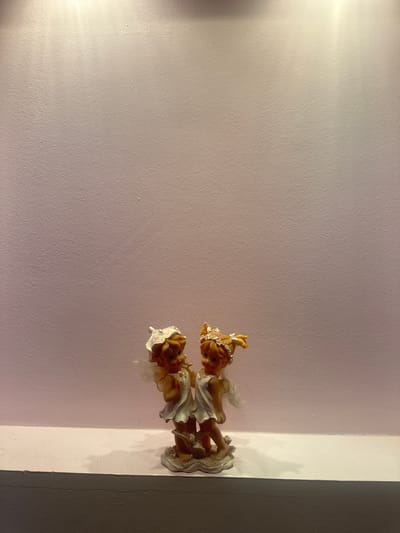 The image features two small figurines resembling angels with blonde hair and light-colored dresses. They are positioned on a surface that appears to be a shelf or ledge, against a wall. The wall has a textured surface and is painted in a pale pink hue, with a lighter colored vertical band to the right of the figurines, suggesting a corner or a dividing line between different wall sections. A shadow is cast on the right side of the wall. The lighting in the scene creates a soft glow on the wall, particularly on the left side, and highlights the details of the figurines. The scene appears to be indoors, possibly in a home or a display area. The figurines are the main subjects, and no specific activity or event is depicted; they are presented as decorative objects. There is no visible text in the image. The overall mood is serene and decorative.