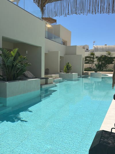 This is a medium-shot, eye-level outdoor photograph of a modern resort swimming pool area on a sunny day. The pool is in the foreground, filled with clear, turquoise water and tiled with light-colored square tiles. The water has ripples and reflections from the surrounding architecture and vegetation.

To the left of the pool, there is a raised, rectangular concrete planter box with a light-colored tile finish. Inside the planter box, a large Bird of Paradise plant with broad green leaves is visible. Behind the planter box, there is a recessed area with a light-colored lounge chair covered with a grey cushion. Adjacent to this, a series of stepped platforms with light-colored tile finishes lead up to another recessed area.

Further back, a multi-story, contemporary building with white and light beige walls is prominent. The building has clean lines and sharp angles. Balconies with glass railings are visible on the upper levels. There are also recessed architectural elements that appear to be part of the rooms or suites.

In the background, there are more buildings with white facades and some vegetation, including trees and climbing plants with green leaves and some pink flowers. A satellite dish is visible on one of the rooftops.

The sky is a clear, bright blue. A decorative element made of hanging ropes is visible at the top of the frame, casting shadows. The overall aesthetic is minimalist and luxurious, suggesting a high-end vacation destination. The scene is devoid of people. This appears to be a hotel or villa complex, likely located in a sunny, coastal region. Given the architectural style and the vegetation, it is plausible this is in the Cyclades, such as Santorini, Greece, perhaps in Thira Municipal Unit.