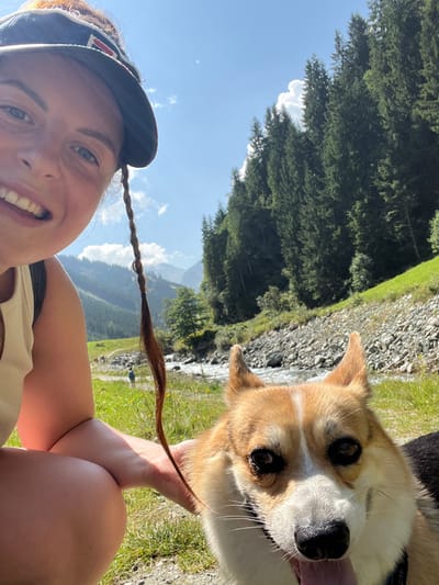 An outdoor, eye-level photograph captures a female human and a Pembroke Welsh Corgi in a mountainous valley setting identified as Reitlehen, Austria. The female subject, partially visible in the left foreground, wears a dark baseball cap and a light top, smiling towards the camera. A long, braided strand of brown hair extends vertically from her head. Her right hand is positioned near the dog's head. The Corgi, a tri-color (fawn, white, and black) dog with erect ears, occupies the right foreground, looking towards the camera with its tongue slightly extended. The immediate ground is covered in green grass with sparse yellow flora. Behind the subjects, a rocky riverbed with flowing water is visible. Across the river, a dirt path extends, with two distant human figures present. The background features steep, conifer-forested mountain slopes rising on the right, and additional distant mountains in the center. The sky is clear blue with scattered white cumulus clouds, indicating bright daylight conditions.