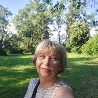 The image is a portrait of a woman taken outdoors, likely in a park or a grassy area with trees. The woman has blonde hair and is looking towards the camera. She is smiling slightly, and her expression appears relaxed and happy. She is wearing a striped top with straps that go over her shoulders. The background is a blur of green trees and grass, with sunlight dappling through the leaves, suggesting it is daytime and the weather is pleasant. The image is taken from a low angle, with the woman's face filling a significant portion of the frame. The orientation of the image is landscape, but the subject is positioned such that her head is towards the left side of the frame. There is no visible text in the image.