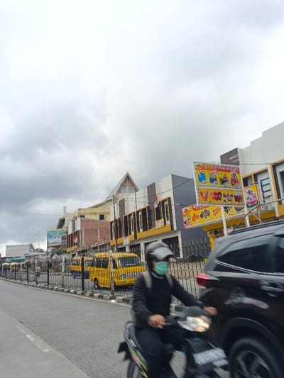 The image captures a street scene in Palayan City, Philippines, under an overcast sky with dark grey clouds. In the foreground, a person wearing a black long-sleeved shirt, dark pants, a grey full-face helmet with the visor raised, and a light blue surgical mask is operating a dark-colored motorcycle. A dark backpack is visible on the rider's back. The motorcycle's headlight is illuminated, and motion blur suggests forward movement. A dark-colored SUV is partially visible to the right of the motorcycle, also appearing to be in motion.
The right side of the street is bordered by a black metal fence, behind which are multiple multi-story commercial buildings. These structures display facades in white, grey, brown, and yellow. One building features a distinctive gabled roofline. Red and white banners are visible on the exterior of several buildings. A prominent yellow sign for "GLOBAL BAKERY" is displayed, showcasing images of cakes, ice cream, and other baked goods. Below it, a smaller yellow sign presents various snack items. Several yellow vans or utility vehicles are parked or stationary behind the fence. The asphalt road surface includes a white solid line on its left edge. Utility poles, electrical wires, and distant billboards are visible on the left side of the frame.