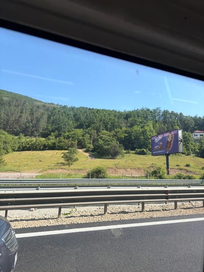 The image captures a roadside view from inside a vehicle, looking out towards a hilly, tree-covered landscape under a clear blue sky. A highway guardrail runs horizontally across the foreground, separating the road from a grassy embankment dotted with bushes and a dirt path. In the middle ground, a billboard advertising "Milka" ice cream is prominently displayed against the backdrop of the lush green hillside. A small building is visible on the right side of the hill. The road surface itself is dark asphalt with a white lane marking. The scene appears to be in a rural area, possibly on the outskirts of Churek, Bulgaria, given the natural scenery and the presence of a highway.