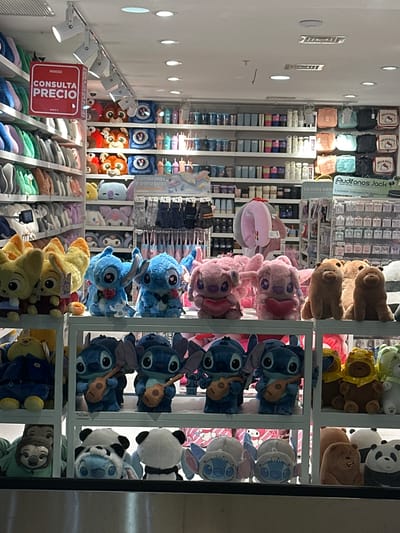The image shows a store shelf in Pudahuel, Chile filled with various stuffed toys. There are pink, blue and yellow plush animals in the central shelves. The back shelves contain various merchandise including bottles and luggage. A red sign displays the word "PRECIO" and the Miniso logo. The image appears to be taken from inside the store, looking toward the shelves. The ceiling has bright track lighting.