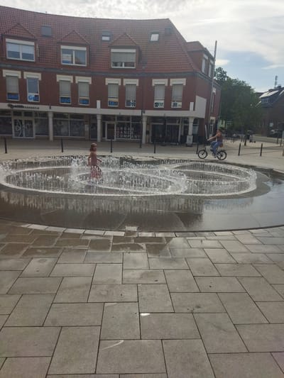 This is a full-shot, eye-level, outdoor photograph of a public square in Monheim am Rhein, Germany, featuring a circular fountain with multiple jets spraying water. A young girl in a light-colored dress is standing in the fountain, her body partially obscured by the water. To the right of the fountain, a woman is riding a bicycle. In the background, there is a brick building with multiple stories and windows, and a sign that reads "URBAN DANCE COMPANY". The ground is paved with large, rectangular stone tiles. The sky is overcast with some patches of blue.