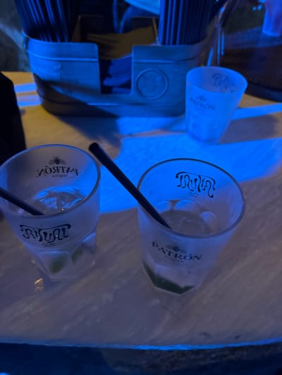 The scene, set in Baja Sardinia, Italy, is illuminated by an intense blue ambient light, suggesting a nighttime or club environment. In the immediate foreground, two frosted, multi-faceted glasses rest on a light-colored, textured surface. The left glass, hexagonal in shape, contains ice cubes, a clear liquid, and two visible lime wedges, with a black straw inserted. Its inner surface displays a mirrored and inverted "PATRÓN TEQUILA" logo, featuring a bee emblem above "PATRÓN," while its outer surface presents a legible "RITUAL CLUB" logo in stylized script. The right glass, octagonal in form, also contains ice cubes, a clear liquid, two lime wedges, and a black straw. Its inner surface exhibits an inverted stylized "m" or wave pattern with "CLUB" below, while its outer surface clearly shows the "PATRÓN TEQUILA" logo with the bee emblem.
Further back on the right, a smaller, empty frosted shot glass is visible, featuring the "PATRÓN" logo and a stylized 'm' or wave graphic near its rim. In the background, a boat-shaped container, appearing to be crafted from wood or metal, is filled with numerous dark drinking straws. This container also displays a circular decorative medallion on its right side. No individuals are present in the image; the focus is solely on the objects and the blue-lit environment.