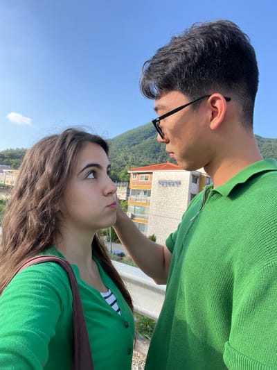 This is a selfie taken outdoors in Dongmyeong-myeon, South Korea, featuring two young people, a man and a woman, looking at each other. The man is wearing glasses and a green polo shirt, while the woman is wearing a green cardigan over a striped shirt. They appear to be in a candid moment, possibly during a date or outing, with a shared look of affection or deep conversation.
The background shows a bright blue sky with a few wispy clouds, indicating it's daytime with good weather. Behind the couple, there's a building with yellow and white siding, possibly a hotel or guesthouse, with a Korean sign on its facade. Further in the distance, a lush green mountainside slopes down, typical of the South Korean landscape. The lighting suggests it's a sunny day, casting soft shadows. The overall mood is intimate and romantic, with the couple engrossed in each other. The image is oriented vertically, with the subjects close-up and filling most of the frame.