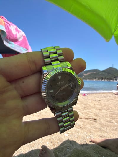 The image shows a hand holding a silver watch at a beach in Nea Irakleitsa, Greece. The watch has a dark dial with Roman numerals and text that reads "SEIKO" and "DAY-DATE". The background is a sunny beach scene with the sea and a hill in the distance. People can be seen in the water and on the sand. A bright green umbrella is partially visible in the upper left corner, and a pink and white patterned towel is on the lower left. The lighting suggests it is daytime, likely mid-morning or afternoon, with clear blue skies and warm weather. The overall mood is relaxed and leisurely, indicative of a vacation or a day at the beach.