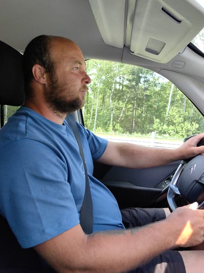 The image captures a male driver operating a vehicle in Негарэльскі сельскі Савет, Belarus, during daylight. The man, with a receding hairline and a full beard, is seated in the driver's position, wearing a blue short-sleeved t-shirt and dark shorts. A black seatbelt is visible across his chest and lap. He is looking forward and slightly to the right, out the front windshield. His left hand grips the black steering wheel, which has a silver-colored spoke and a discernible Citroën emblem. Sunlight illuminates his right forearm and hand, also positioned on the steering wheel. The vehicle's interior features a light grey headliner, a light grey sun visor with a central opening, and a light grey A-pillar marked with "AIRBAG." The left door panel, visible below his arm, is dark and contains window control buttons. Outside the vehicle, a dense treeline of green deciduous trees is visible, indicative of a forested environment. A white roadside barrier or guardrail runs parallel to the road.