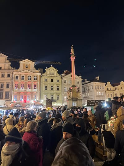 The image depicts a crowded nighttime scene in Old Town Square (Staroměstské náměstí), Praha 1, Czechia. Hundreds of individuals, predominantly adults, are densely packed in the lower two-thirds of the frame, dressed in winter coats, hats, and scarves. Their faces are largely obscured or partially visible.

In the midground, various market stalls or temporary structures, adorned with string lights and greenery, are visible among the crowd. A prominent white sign in the mid-right advertises "PEČENÉ KAŠTANY" (Roasted Chestnuts) and "ROASTED NUTS."

The background features a row of multi-story historic buildings under a dark night sky. The central building has an orange-brown facade and is extensively decorated with red and white string lights, wreaths, and figures resembling Santa Claus in its windows. To the right of these buildings stands the Marian Column, a tall, ornate stone column topped with a statue, likely of the Virgin Mary, illuminated with a reddish-pink light. A golden halo is visible around the statue's head.

Above the buildings in the upper left, the yellow boom of a construction crane is silhouetted against the dark sky. The sky itself is dark, with several small, blurry green light artifacts or lens flares visible in the upper right quadrant. Artificial lighting from buildings, market stalls, and the Marian Column illuminates the scene.