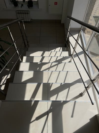 The image displays an unoccupied interior staircase within a building in Enem, Russia. The perspective is a high-angle view looking down a flight of approximately nine light-colored, beige-tiled steps, each featuring a metallic anti-slip strip along its edge. Polished stainless steel railings with multiple horizontal bars flank both sides of the staircase. Intense sunlight streams from a large window on the right, casting distinct, elongated diagonal shadows of the railings across the steps and the tiled floor below. To the left of the stairs, an interior wall is visible, featuring a dark potted plant on a ledge above a white radiator. In the upper background, a white wall holds a mounted white cabinet with a red "i" symbol, indicating a fire safety utility box. Through the large window on the right, an exterior paved area, constructed from light-colored rectangular blocks, is visible. No individuals, actions, or interactions are present in the scene.