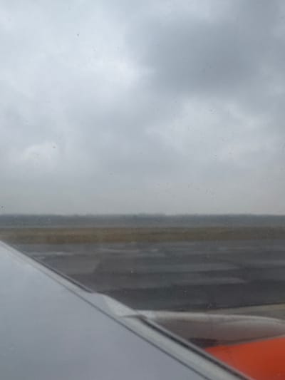The image presents a forensic view from inside an aircraft at Milan Malpensa Airport (MXP) in Somma Lombardo, Italy. The lower left quadrant displays a portion of a grey and white aircraft wing. Below the wing's trailing edge, in the bottom right, an orange component, likely part of an engine cowling, is visible. The midground consists of a dark grey asphalt or concrete runway surface, appearing wet and reflective, suggesting recent precipitation. Adjacent to the runway is a strip of muted brown, possibly dry ground or vegetation. The horizon line is indistinct, with a faint, hazy line of dark, blurred forms in the far background, indicative of distant landscape or structures. The sky is entirely overcast, dominated by uniform grey clouds, indicating low visibility conditions. Small water droplets or specks are scattered across the aircraft windowpane. No people are visible. The scene captures the static presence of an aircraft on the tarmac under a cloudy, possibly rainy, sky.