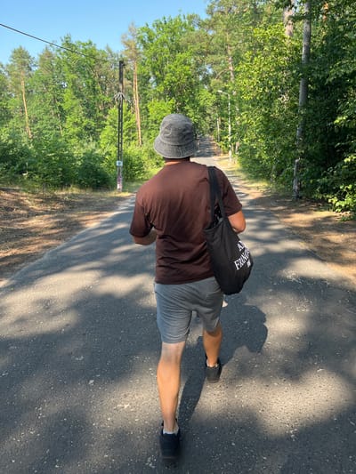 A person is walking on a paved path outdoors. The person is wearing a brown t-shirt, grey shorts, a grey bucket hat, and black shoes with white socks. They are carrying a black duffel bag over their right shoulder. The bag has white text printed on it. The scene is a path lined with trees and foliage. The path is paved and appears to be asphalt with visible stones. Sunlight casts shadows on the path, suggesting it is daytime. The sky is visible and appears clear. There is a utility pole with wires on the left side of the path. The overall setting appears to be a park or a rural area. The text on the bag reads "ALEXANDRIA" in large letters and "FRANCESCO" in smaller letters below it.