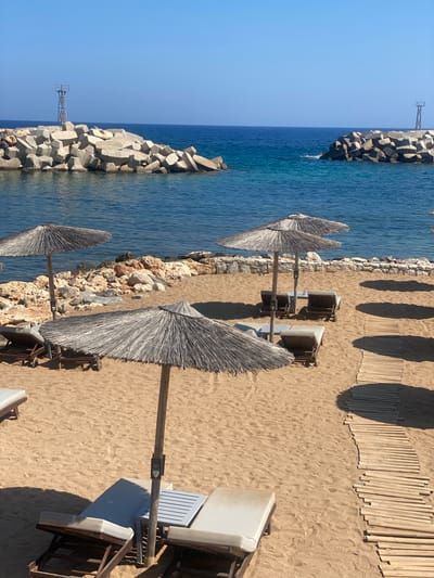 This is a high-angle, eye-level shot of a sandy beach in Panormos, Greece, on a clear, sunny day. The beach is dotted with thatched umbrellas and empty lounge chairs. A wooden walkway leads across the sand. In the background, the turquoise waters of the Aegean Sea meet a rocky breakwater, with a small lighthouse-like structure on one end. The scene conveys a sense of tranquil, unoccupied leisure.