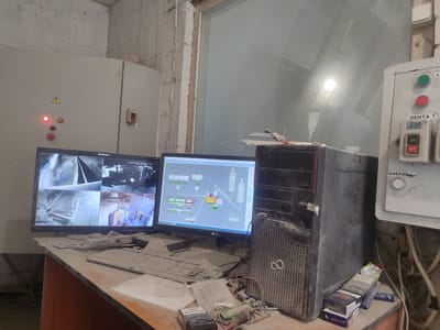The image depicts a control room in Bilohorodka, Ukraine, likely part of an industrial or processing facility. The scene is cluttered and dusty, with equipment covered in a layer of fine particulate matter.
Dominating the foreground is a desk with two computer monitors, a keyboard, and various objects including several packs of cigarettes, a screwdriver, and other miscellaneous items. The left monitor displays a multi-camera feed, showing different views of what appears to be a bulk material handling or processing operation. The right monitor shows a schematic diagram of a process plant, with pipes, silos, and equipment represented by icons. A large, dusty computer tower sits to the right of the monitors.
Behind the desk, a large electrical control panel is visible. This panel has several buttons and indicators, including a red and green button labeled "ПУСК ДВИГАТЕЛЯ 1" (Engine Start 1) and a section labeled "ЛЕНТА 1" (Belt 1) with an ON/OFF switch. There is also a button labeled "ВИБРАЦИЯ" (Vibration). To the left of the desk is a tall, gray electrical cabinet with warning symbols and indicator lights, one of which is illuminated red.
The environment appears to be an indoor space with rough concrete walls and a large window or glass partition, through which some light enters, reflecting off the surfaces. The overall impression is one of an operational, albeit somewhat neglected, control station within an industrial setting. No people are present in the image.